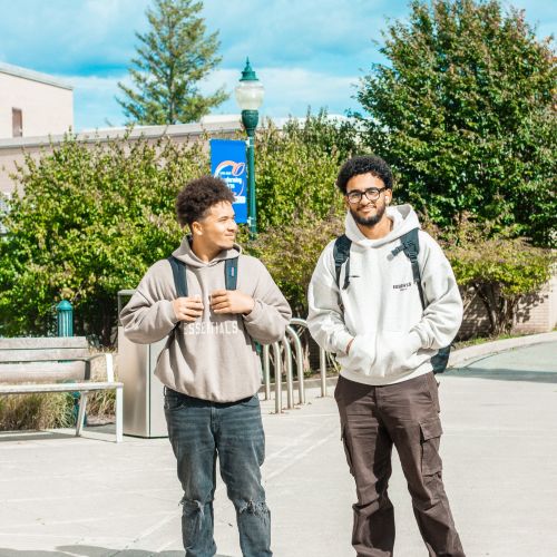 Two male students looking into camera and smiling. There is a SUNY Orange flag in the background.