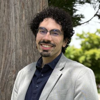 Photograph of Carlos Lassalle standing outdoors in front of a large tree and greenery. Carlos is wearing a light gray blazer over a dark blue shirt with a subtle pattern.