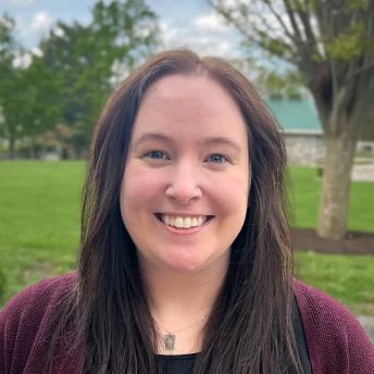 Photograph of Victoria Lucas with long dark hair wearing a maroon sweater and a necklace, standing outdoors in a park-like setting with green grass, trees, and a partly cloudy sky. Background includes a tree trunk and a building with a green roof, suggesting a calm, natural environment.