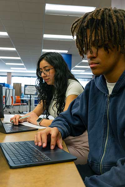 Two students, one male and one female, work at laptops in the SUNY Orange Library